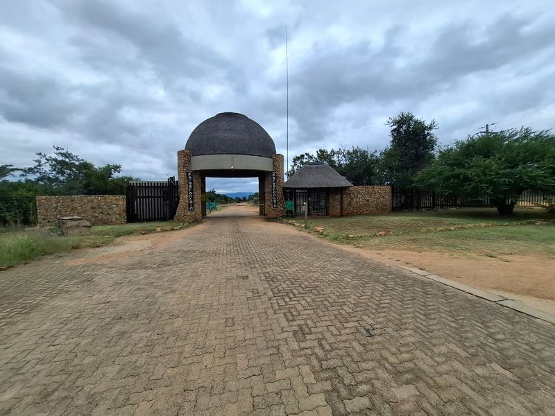 Main entrance gatehouse featuring stone masonry construction, a thatched roof, and a paved driveway leading into the residential estate.