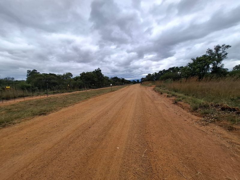 A view of an unpaved access road bordering a property with electric fencing and natural vegetation under a cloudy sky.