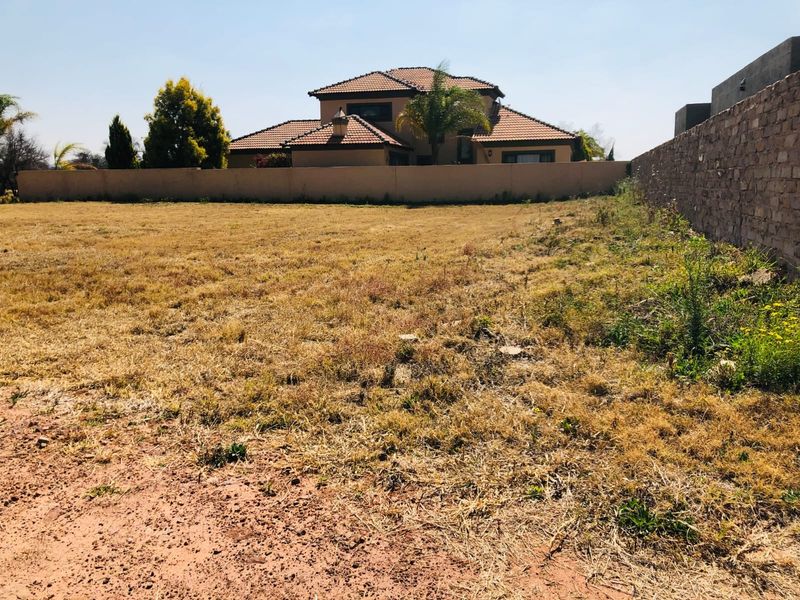 A view of a vacant residential plot featuring dry grass, enclosed by a perimeter wall and a stone boundary wall.