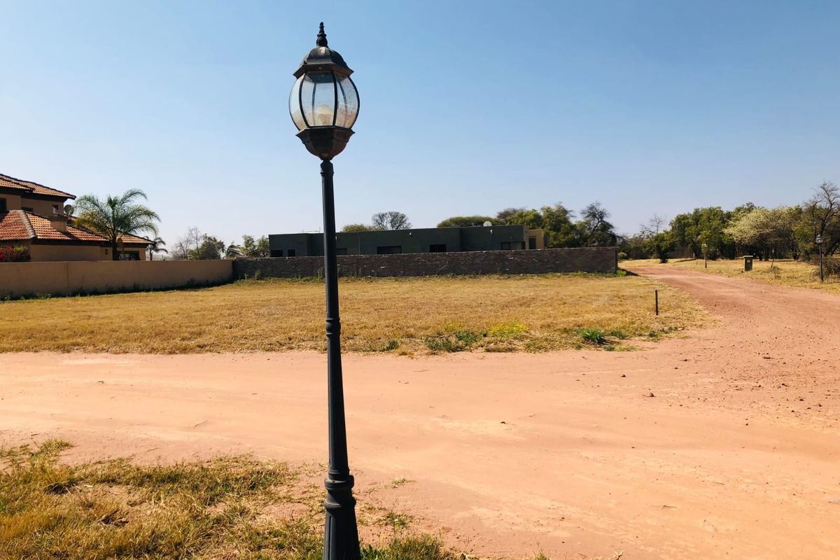 A vacant residential plot featuring a dirt road access, a decorative street lamp, and surrounding boundary walls in a quiet neighborhood setting.