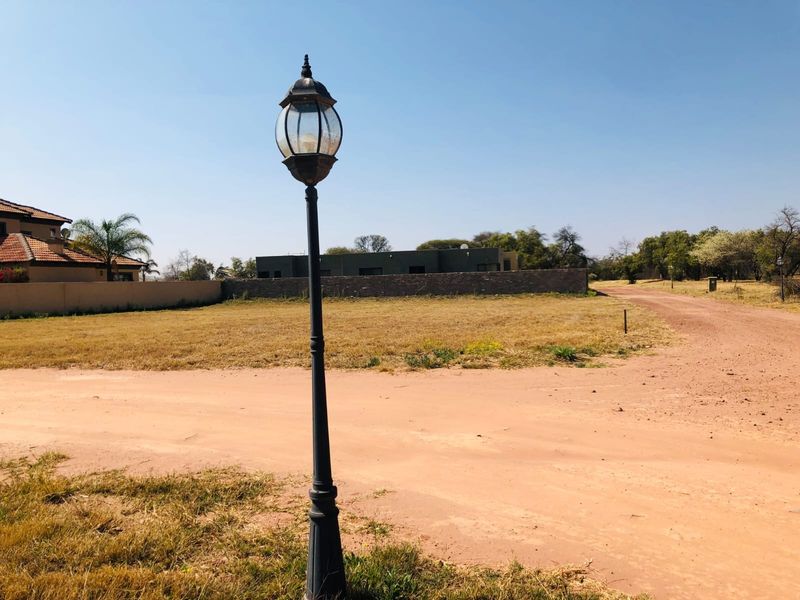 A vacant residential plot featuring a dirt road access, a decorative street lamp, and surrounding boundary walls in a quiet neighborhood setting.