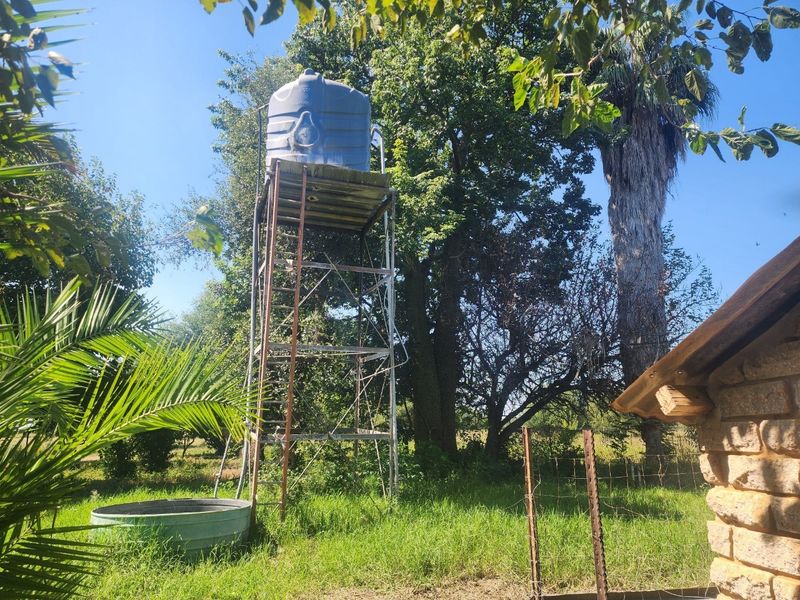Outdoor area featuring a raised water storage tank on a metal stand, accompanied by a metal water trough and perimeter wire fencing.