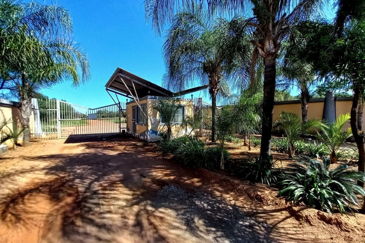 Front entrance view of the property featuring a security gate, guard house, and established palm trees on a dirt access road.