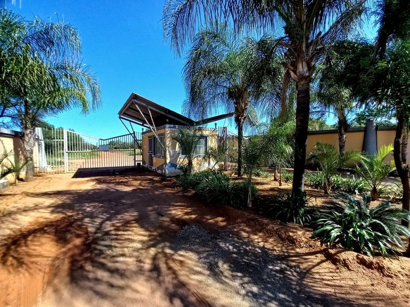 Front entrance view of the property featuring a security gate, guard house, and established palm trees on a dirt access road.