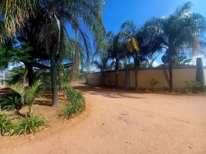 Front view of the residential property featuring a perimeter wall, metal entrance gate, and established garden landscaping along an unpaved driveway.