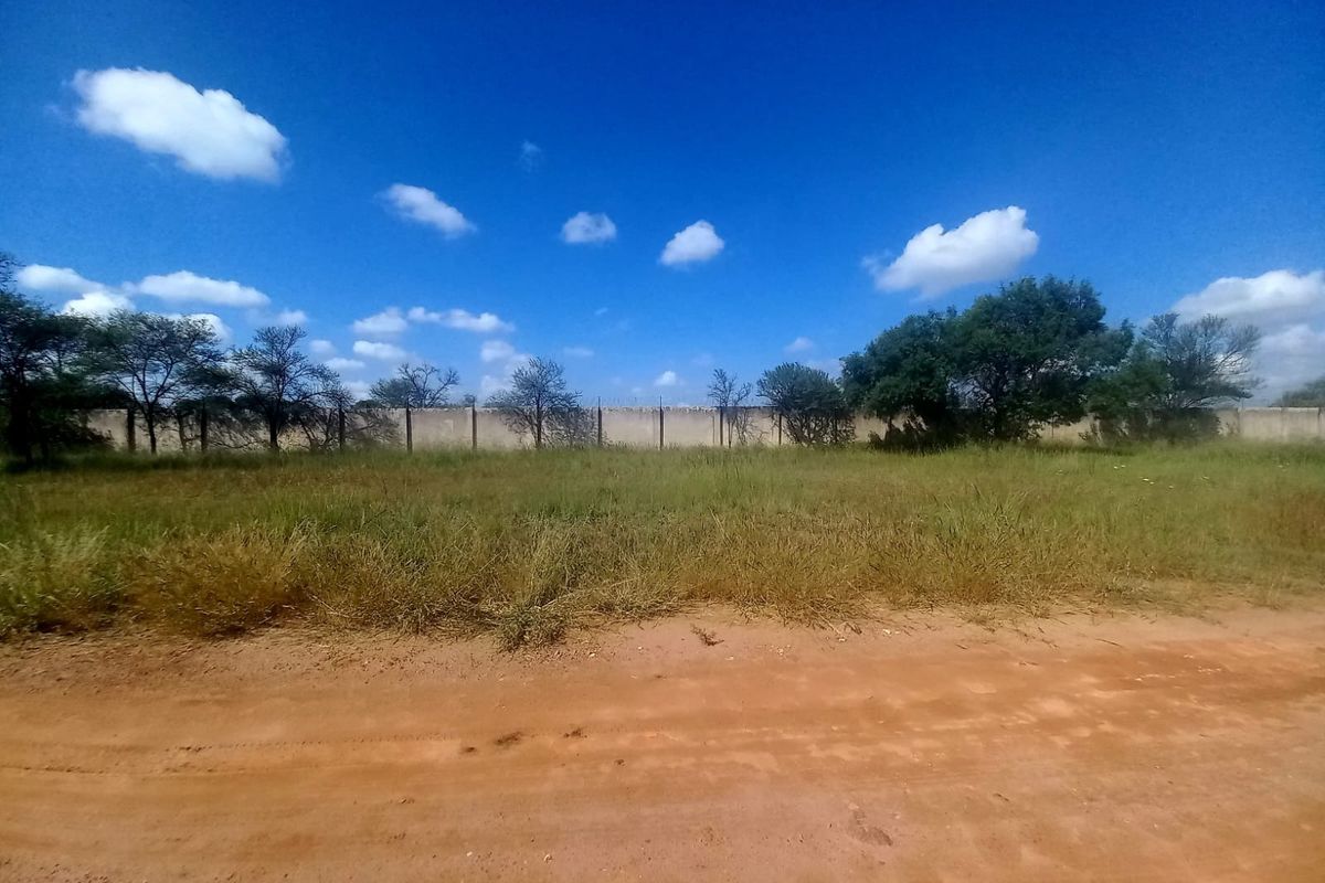 A vacant residential plot featuring natural grass cover, a perimeter wall, and access via an unpaved road.