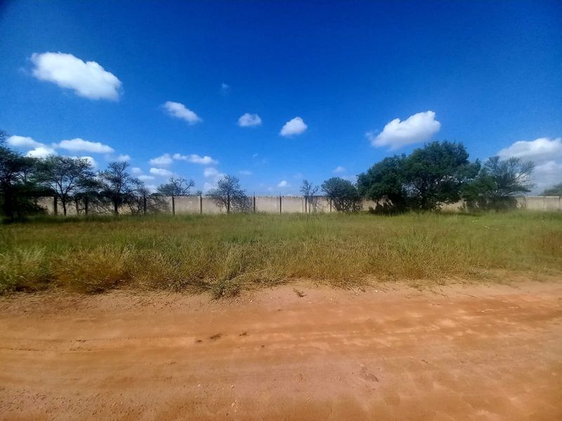 A vacant residential plot featuring natural grass cover, a perimeter wall, and access via an unpaved road.