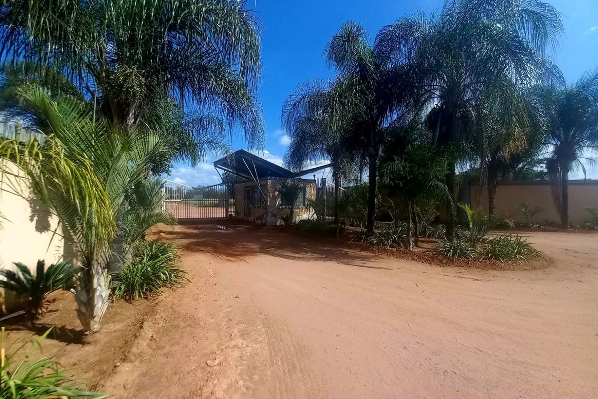 Front entrance view of the property featuring a security gate, guard house, and established palm trees along an unpaved access road.