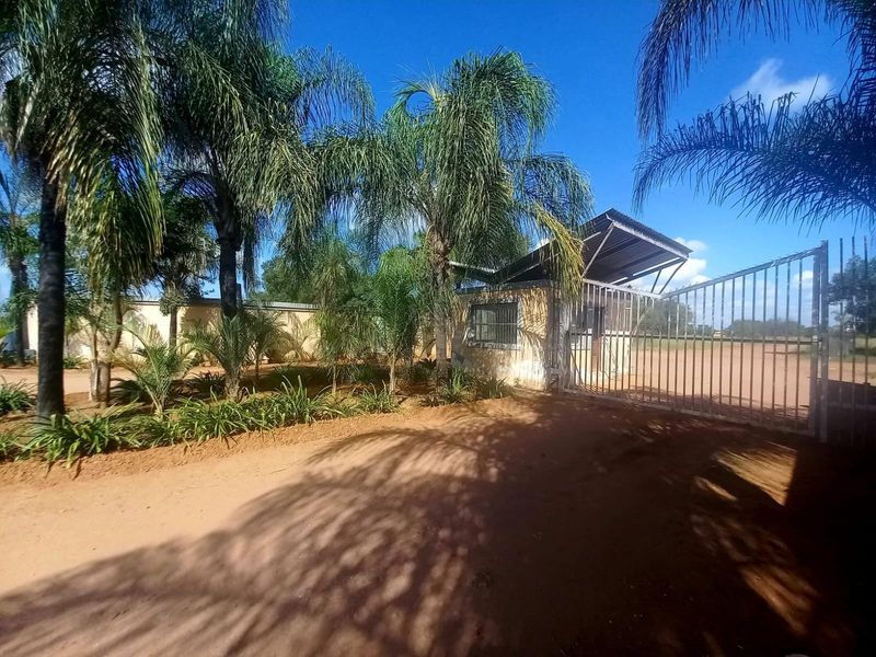 Front entrance view of the property featuring a metal security gate, guard house, and established palm tree landscaping.