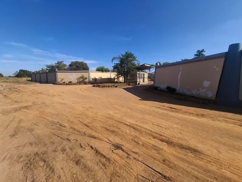 Front view of a residential property featuring a perimeter wall, gated entrance, and an unpaved driveway on a vacant land plot.