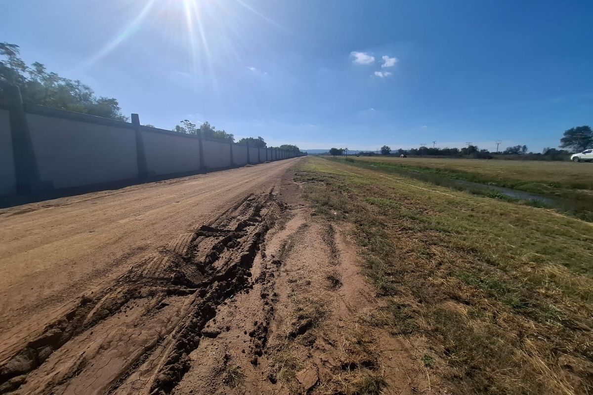 View of the vacant land property featuring an unpaved access road, a long perimeter boundary wall, and adjacent drainage infrastructure.