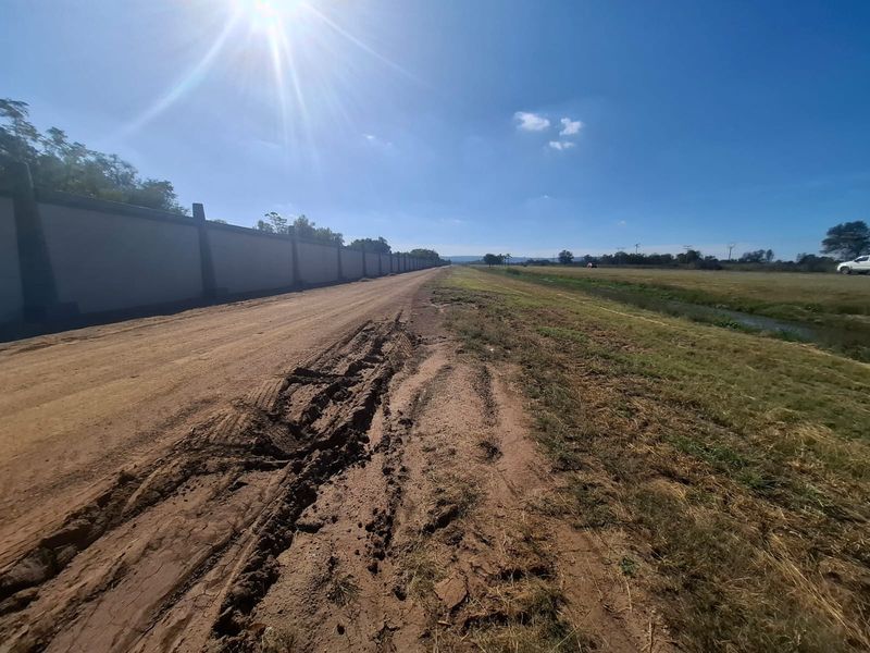 View of the vacant land property featuring an unpaved access road, a long perimeter boundary wall, and adjacent drainage infrastructure.