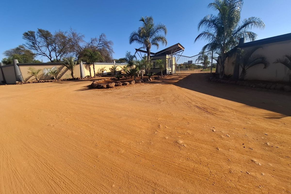 Front entrance view of the property featuring a security gate, guard house, and perimeter wall on a large unpaved residential plot.
