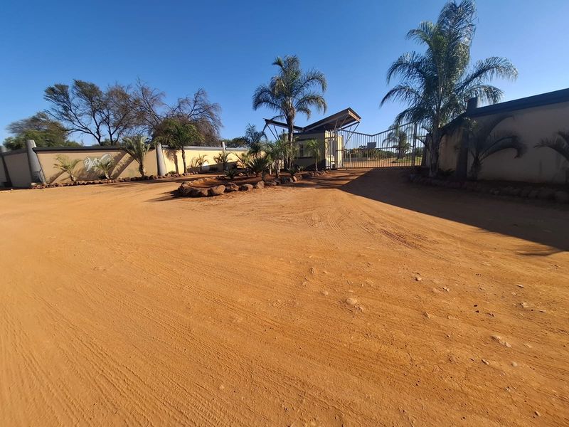 Front entrance view of the property featuring a security gate, guard house, and perimeter wall on a large unpaved residential plot.