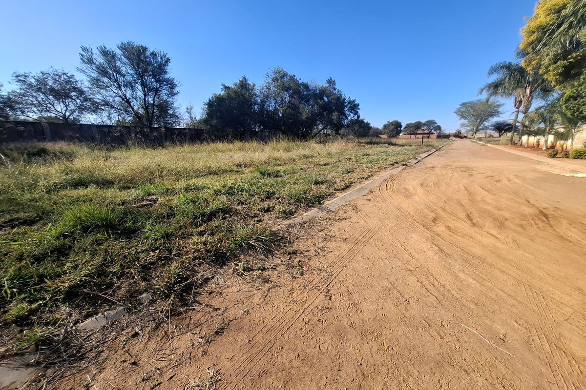 A view of a dirt road adjacent to a vacant residential plot with established perimeter walling and natural vegetation.