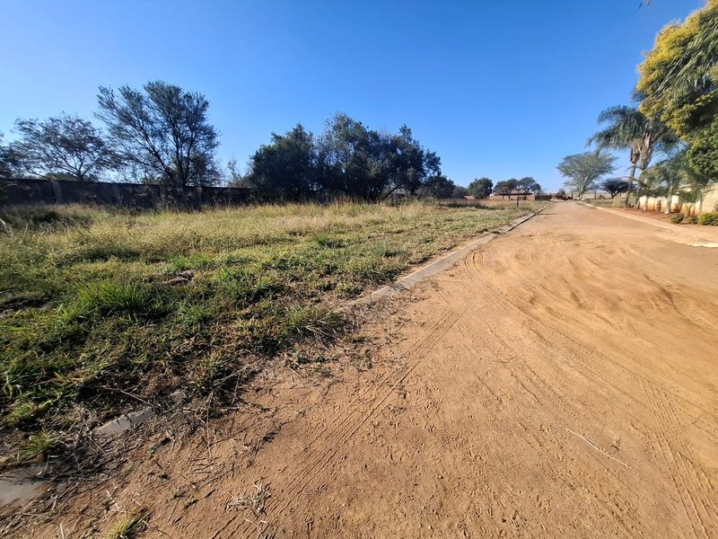 A view of a dirt road adjacent to a vacant residential plot with established perimeter walling and natural vegetation.