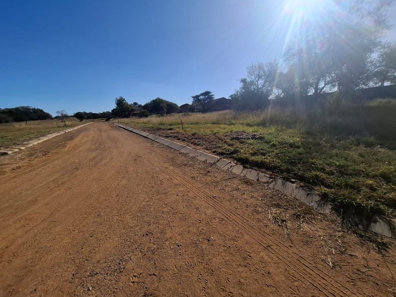 A view of an unpaved residential road featuring established concrete kerbing and surrounding vacant land.