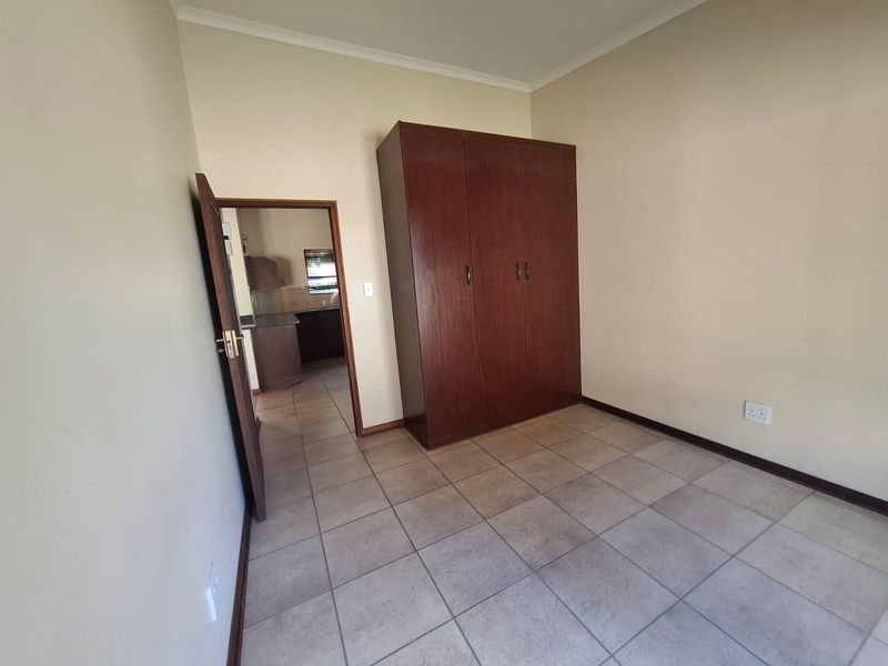 A clean, empty bedroom featuring tiled flooring and a built-in wooden wardrobe, with a view into the adjacent kitchen area.