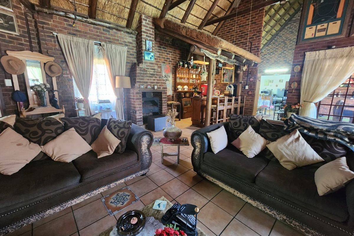 Spacious living room featuring rustic exposed brick walls, a traditional thatched roof with timber beams, tiled flooring, and a built-in fireplace and bar area.