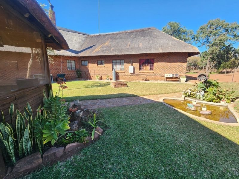 A well-maintained rear garden area featuring a traditional thatch-roofed brick farmhouse, stone pathways, a small garden pond, and a central fire pit.