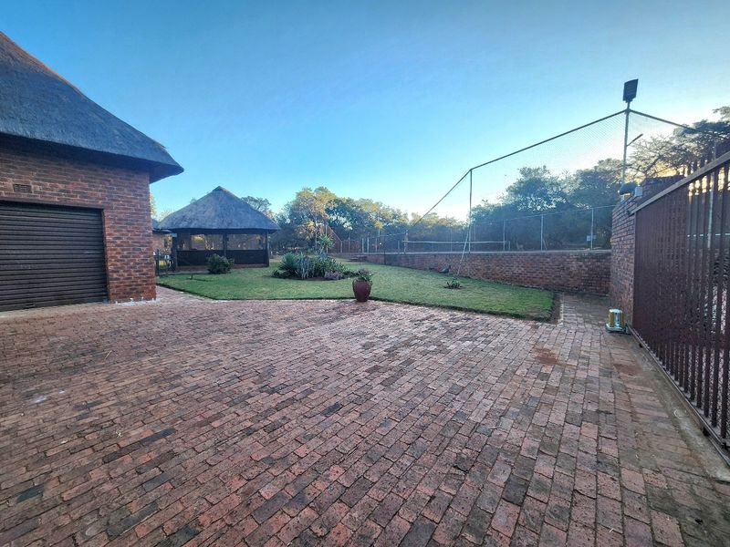 Spacious rear exterior area featuring brick paving, a thatched lapa, and a fenced tennis court, set within a well-maintained farm property.