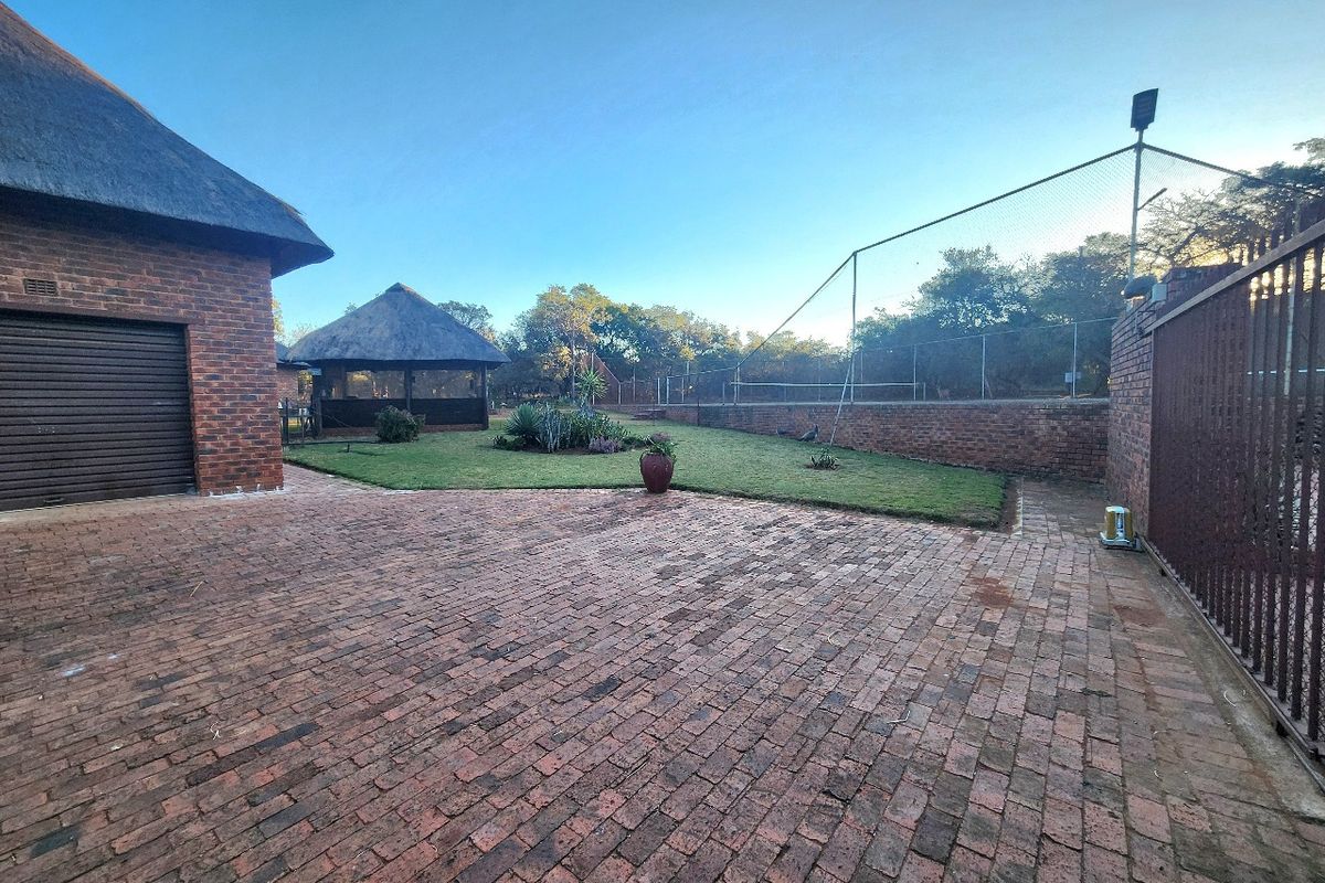 Spacious rear exterior area featuring brick paving, a thatched lapa, and a fenced tennis court, set within a well-maintained farm property.