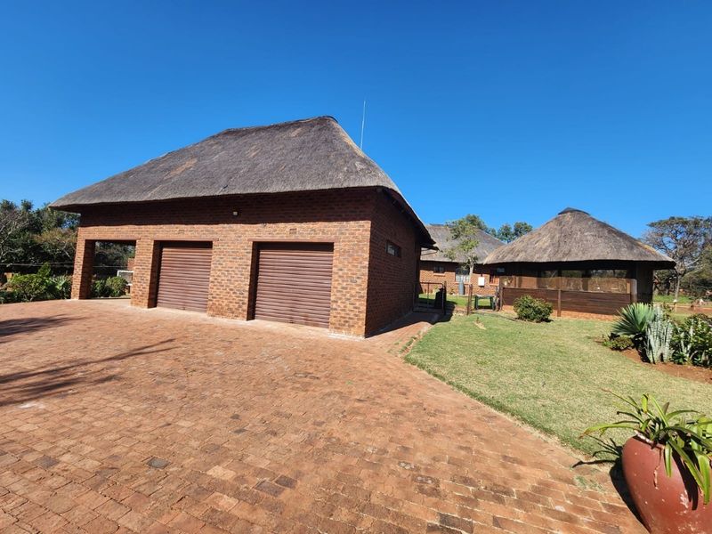 A well-maintained farm property featuring a face brick double garage with a traditional thatch roof, set on a spacious paved driveway with landscaped grounds.