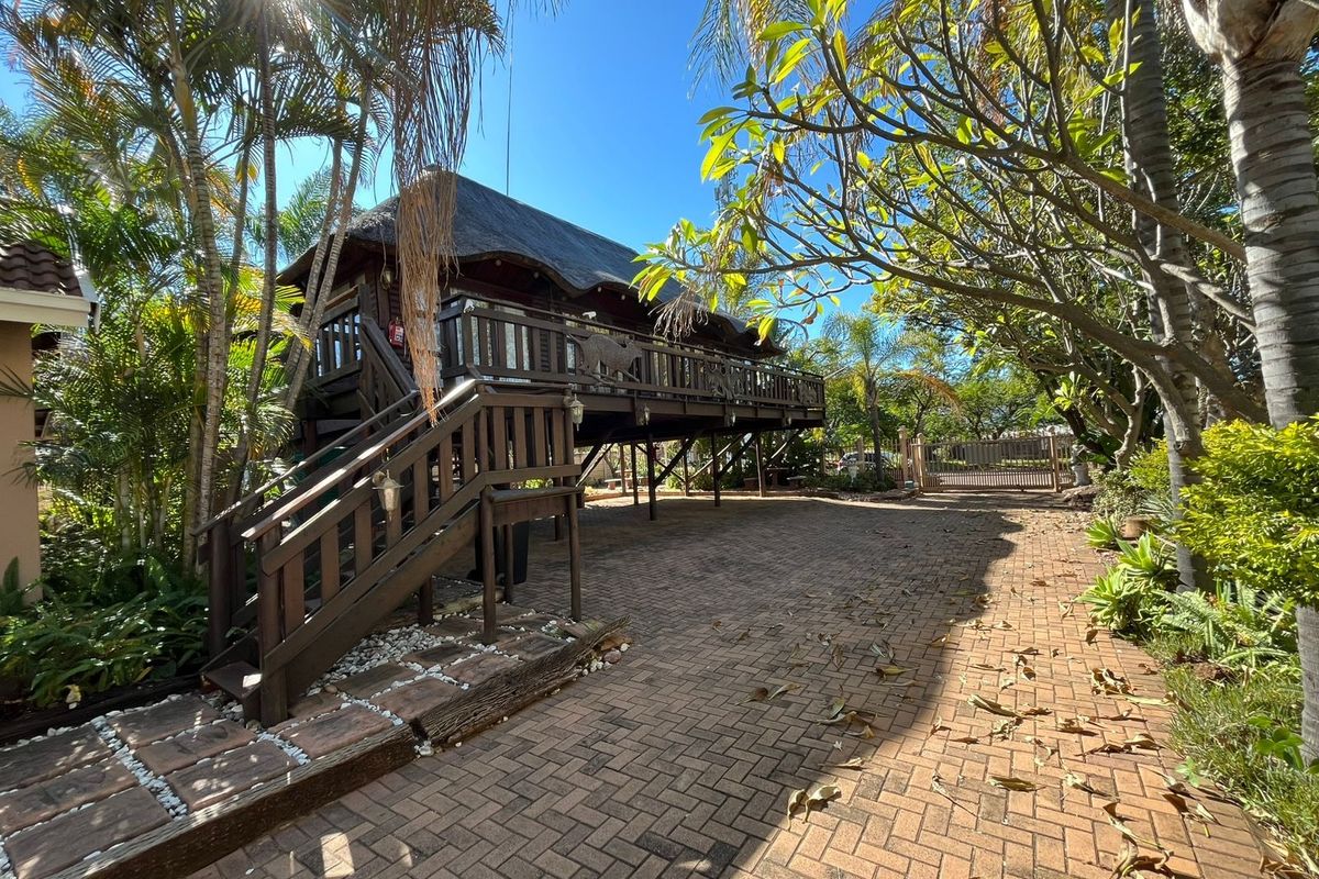 A raised wooden structure with a thatched roof, featuring a deck and staircase, set within a landscaped garden with a brick-paved driveway.