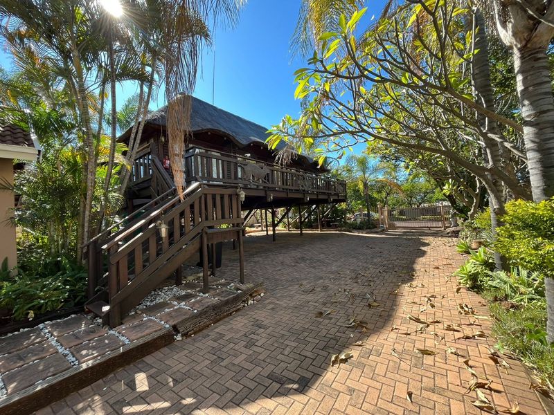 A raised wooden structure with a thatched roof, featuring a deck and staircase, set within a landscaped garden with a brick-paved driveway.