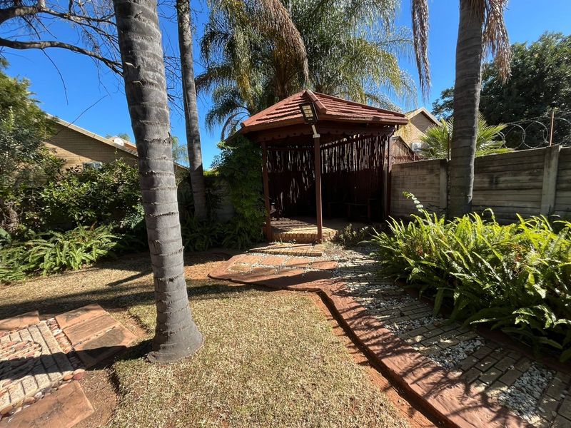 A landscaped garden featuring a wooden gazebo, paved pathways, and mature trees, enclosed by a precast concrete boundary wall with security razor wire.
