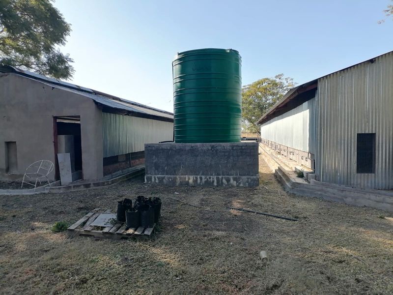 Exterior view of agricultural outbuildings featuring a large green water storage tank mounted on a concrete plinth between two metal-clad structures.