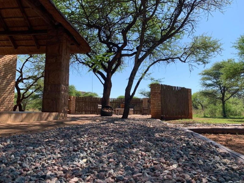 A covered outdoor patio area featuring stone pillars, a thatched roof, and a gravel-surfaced ground, set within a natural bushveld environment.