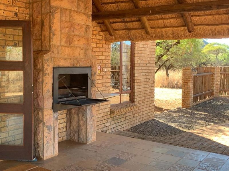 Covered outdoor patio featuring a built-in braai, rustic stone and brick finishes, and a traditional thatched roof overlooking the natural surroundings.