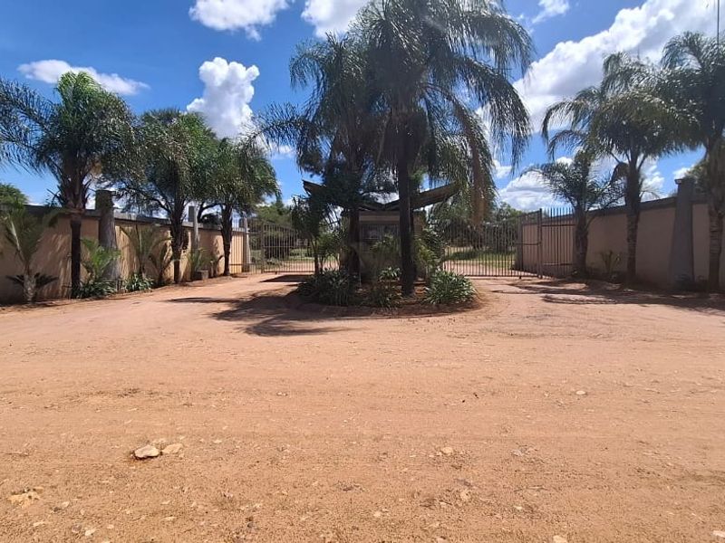Entrance view of a residential property featuring a perimeter wall, security gate, guard house, and established palm trees along an unpaved driveway.