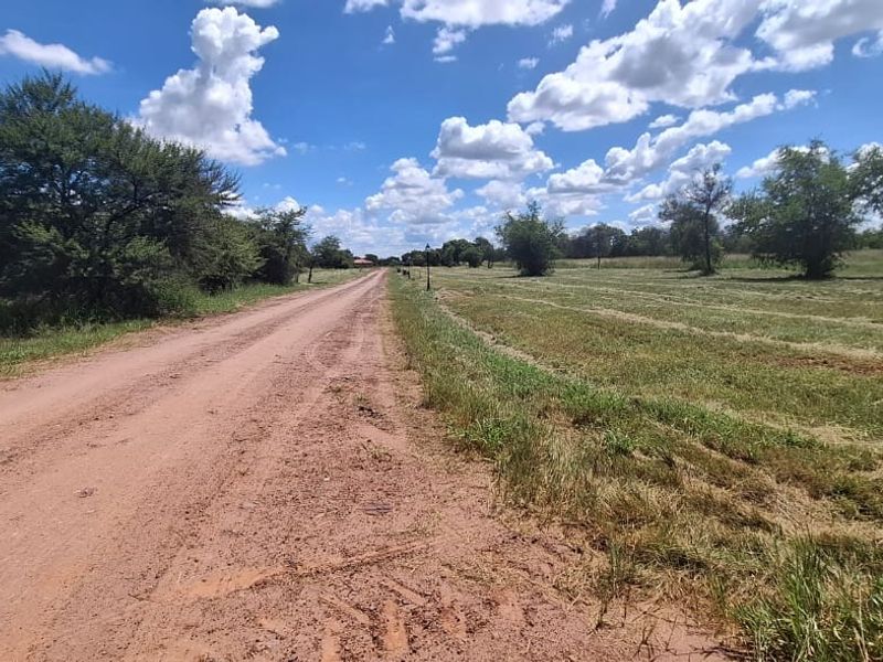 A view of an unpaved dirt road leading through a grassy residential land plot under a clear blue sky.