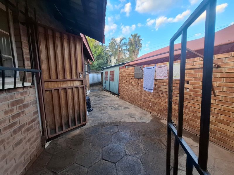 A narrow rear exterior walkway featuring brick boundary walls, paved flooring, and a metal gate entrance.