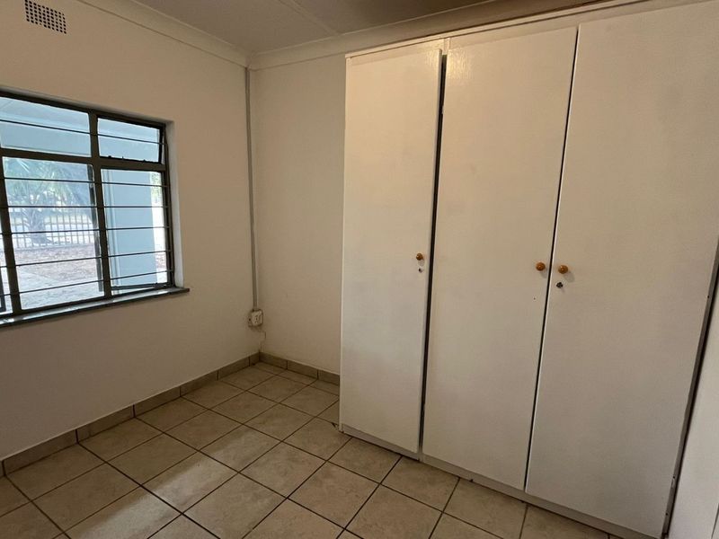 A simple, empty bedroom featuring tiled flooring, built-in cupboards, and a window with security bars.