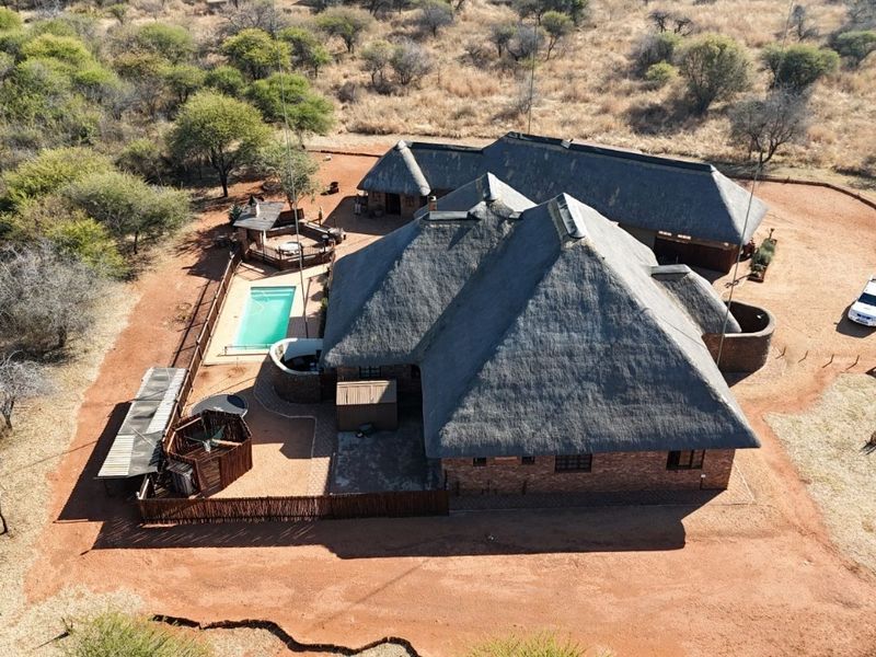 Aerial view of a large thatched-roof residence featuring a swimming pool and outdoor entertainment area set within a natural bushveld landscape.