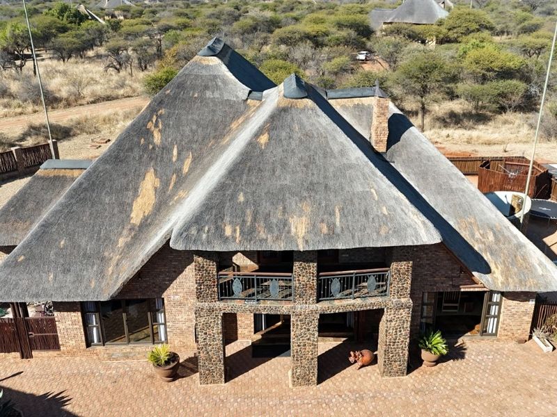 Aerial view of a large house featuring a prominent thatch roof, stone-clad pillars, a balcony, and an outdoor swimming pool in a bushveld setting.