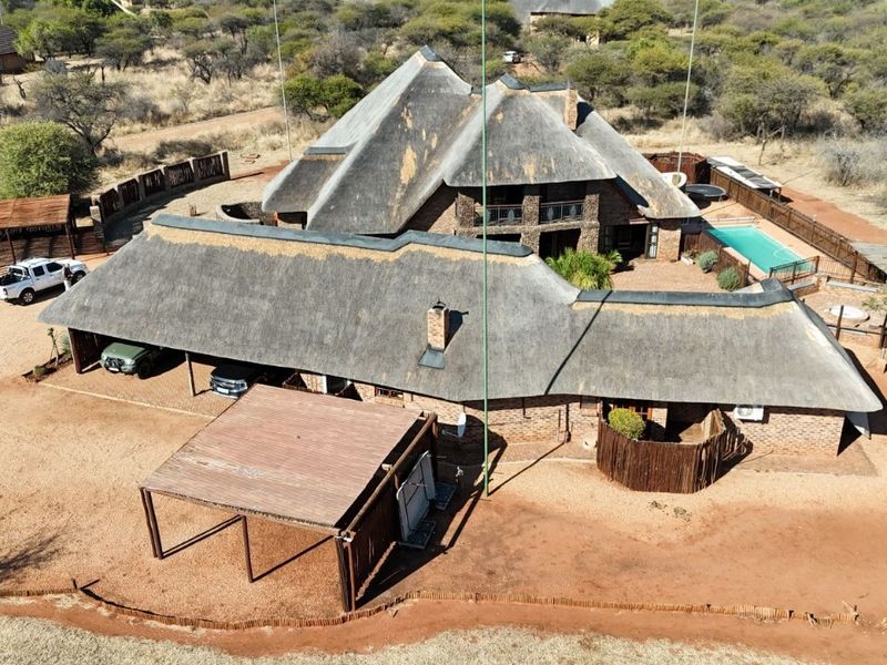 Aerial view of a large thatched-roof house featuring a swimming pool, multiple carports, and expansive surrounding grounds in a bushveld setting.