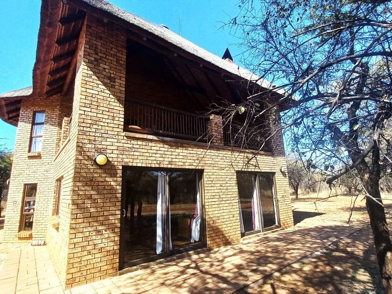 Rear view of a two-story face brick house featuring a traditional thatched roof, an upper-level balcony, and sliding glass doors opening onto a paved patio.