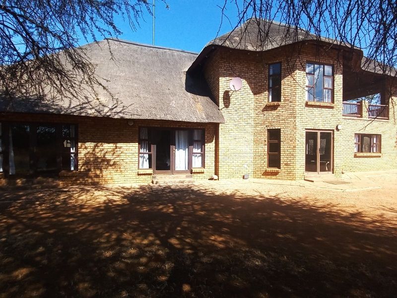 A rear view of a double-story face brick house featuring a traditional thatch roof, balcony, and multiple access points via sliding doors.