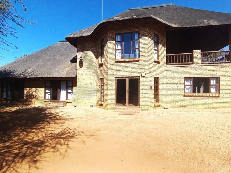 A two-story face brick house featuring a traditional thatch roof, a covered balcony, and multiple access points, set on a natural sandy landscape.