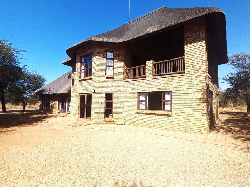 A two-story face brick house featuring a traditional thatch roof, a covered upper-level balcony, and a paved outdoor area set in a natural landscape.