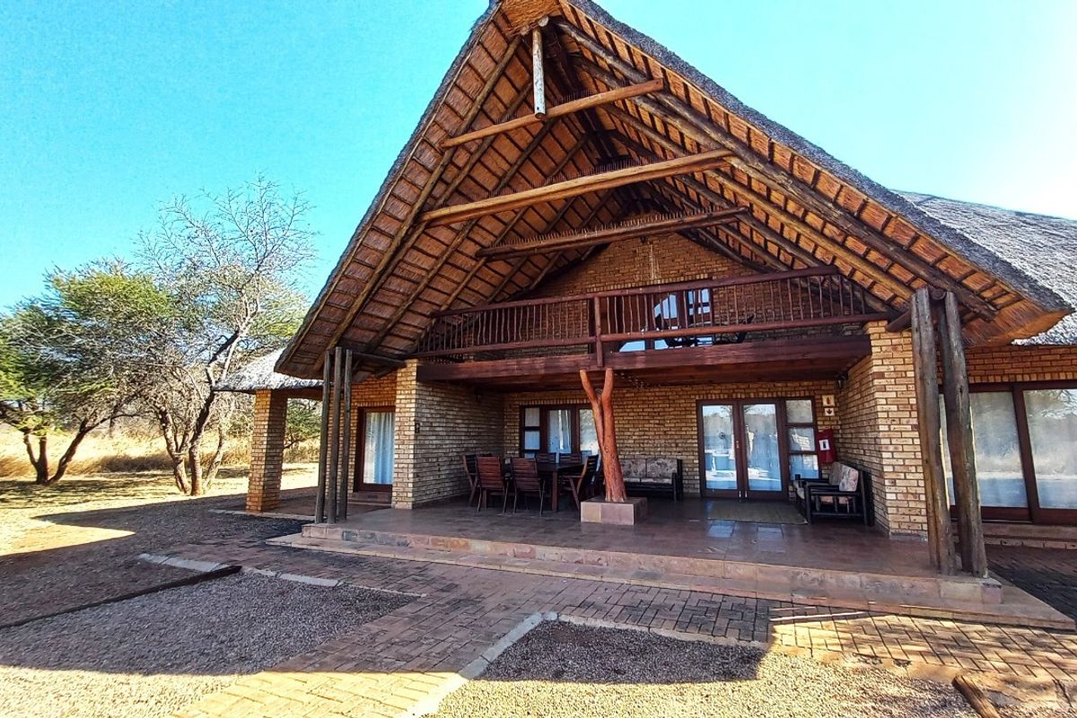A spacious rear exterior view featuring a traditional thatch roof, exposed timber trusses, a covered patio, and a wooden balcony overlooking the surrounding landscape.