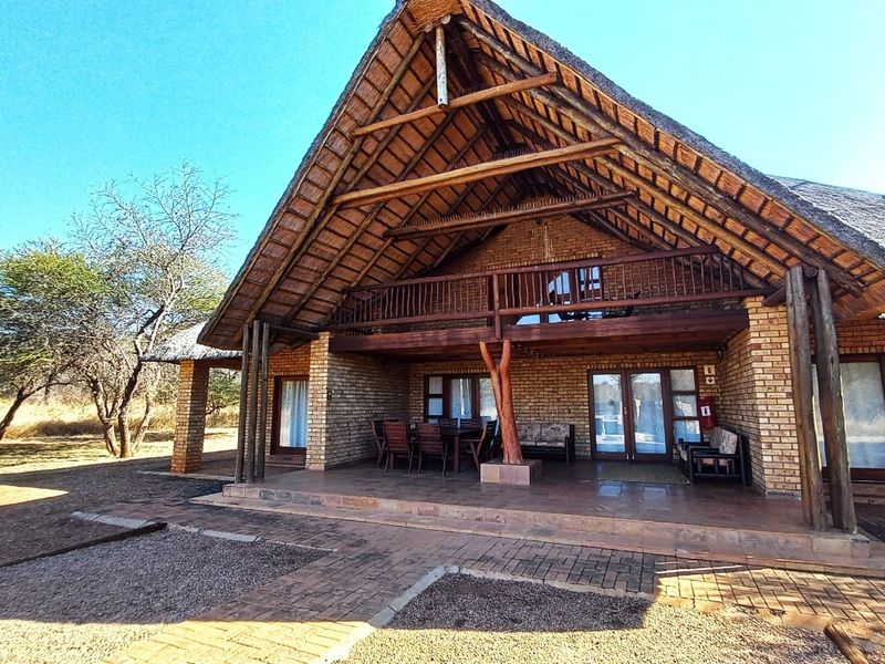 A spacious rear exterior view featuring a traditional thatch roof, exposed timber trusses, a covered patio, and a wooden balcony overlooking the surrounding landscape.