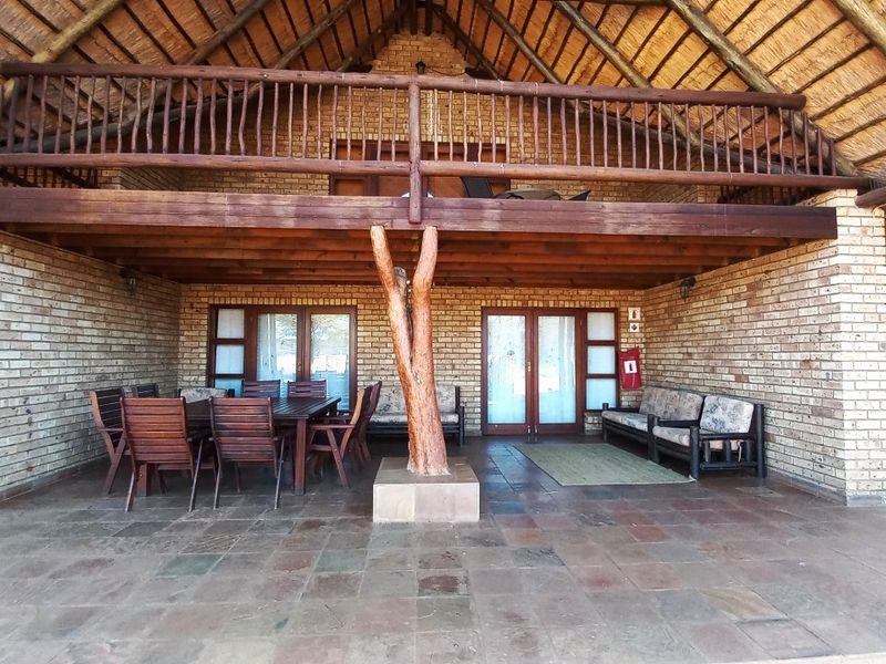 A spacious covered patio featuring a traditional thatched roof, exposed brickwork, stone tile flooring, and a wooden balcony overlooking the outdoor seating area.