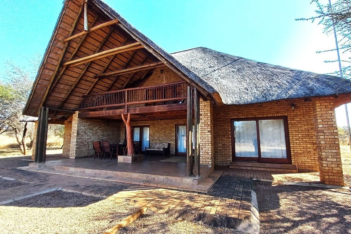 A rear view of a brick house featuring a large thatched roof, a covered patio area, and an upper-level timber balcony.