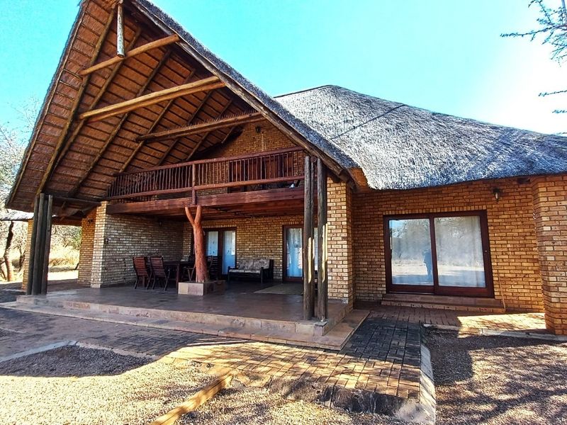 A rear view of a brick house featuring a large thatched roof, a covered patio area, and an upper-level timber balcony.