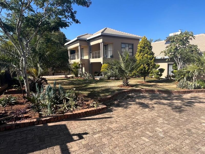 A spacious rear exterior view of a two-story house featuring a paved driveway, landscaped garden, and an upper-level balcony.